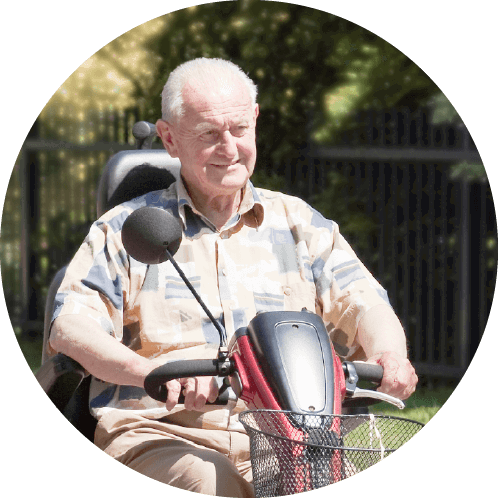 An older adult sits on a mobility scooter in a park-like setting, wearing a patterned shirt and light trousers. The scooter has a front basket and rearview mirror, with greenery and a metal fence in the background.